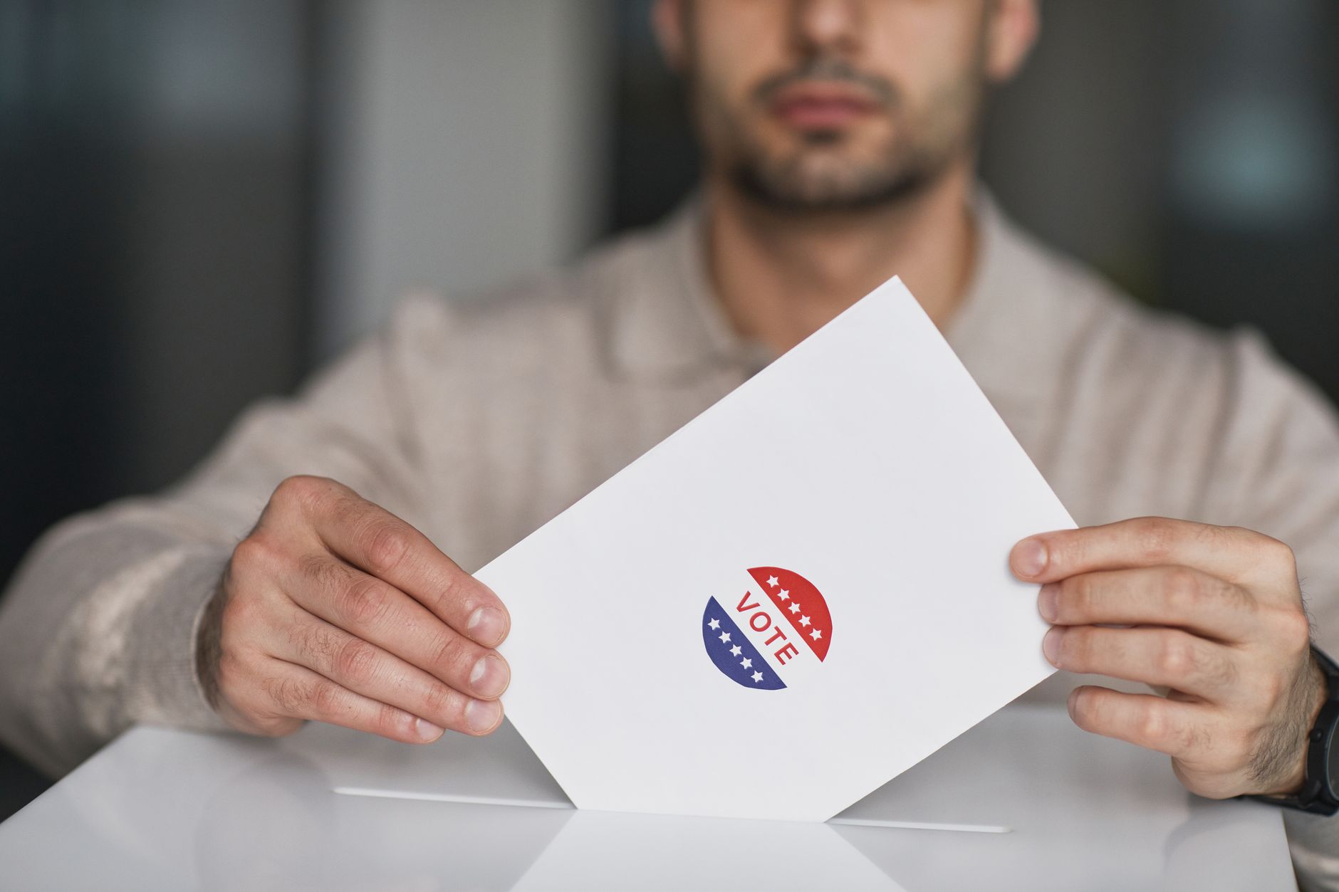 A man holds a white envelope with a red, white and blue "VOTE" sticker over a white box with a slot, as if casting a ballot.