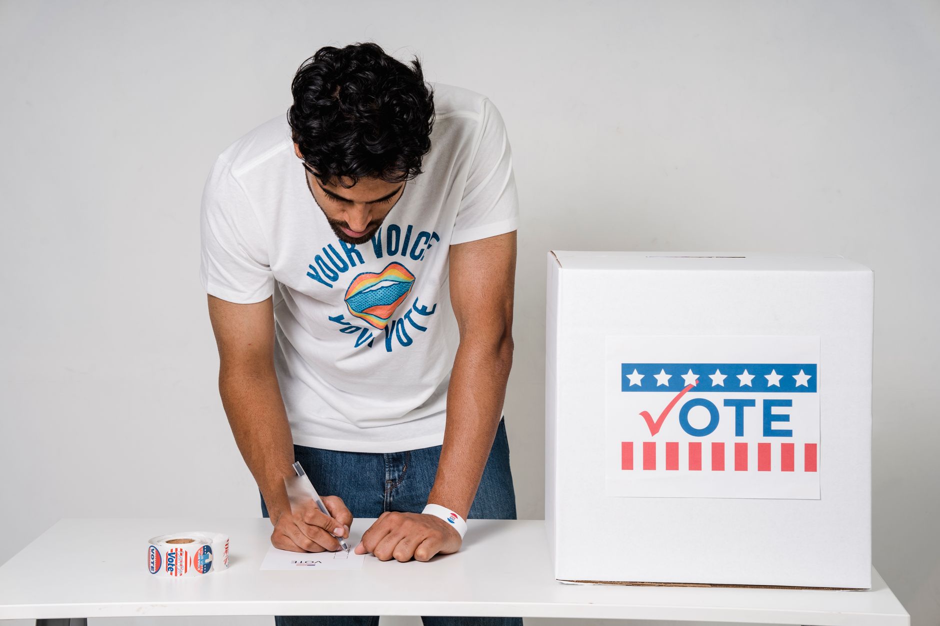 A man standing at a white table filling out a paper ballot beside a ballot box.