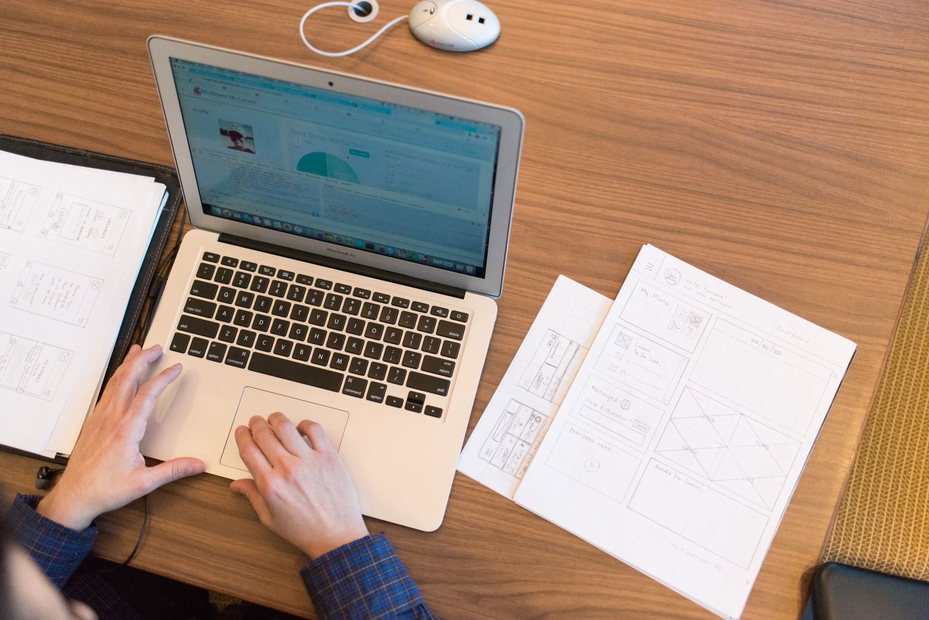 Overhead view of a person working on a laptop at a wooden desk with paper sketches nearby.