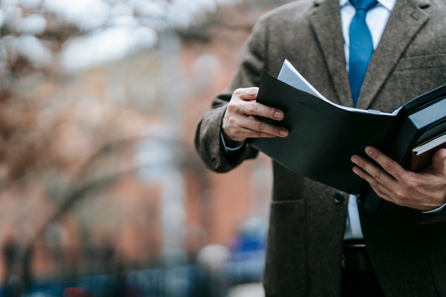 A person wearing a suit jacket and tie reviews documents inside an open black folder outdoors.